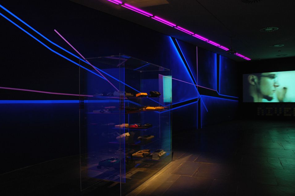 Vitrina de museo con videoconsolas antiguas iluminadas en primer plano y de fondo un muro pintado con pintura flúor y luz negra.